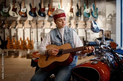 Young female rocker playing guitar at music shop sitting on motorcycle