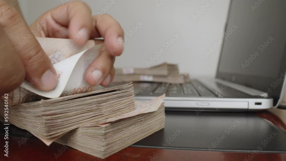 Select focus of hands counting a lot of thousands Thai baht banknotes money near laptop or notebook computer. The baht is the official currency of Thailand.
