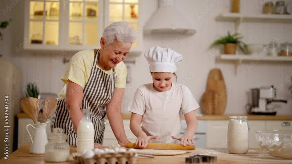 Granny teaching her granddaughter to cook cookies in kitchen at home. Happy family cooks together. Grandma rolls out dough uses rolling pin. Smiling pre-teen girl in cook's cap helps to senior woman. Granny teaching her granddaughter to cook cookies in kitchen at home. Happy family cooks together. Grandma rolls out dough uses rolling pin. Smiling pre-teen girl in cook's cap helps to senior woman.