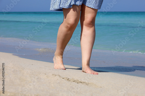 Barefoot woman in short dress walking by sea waves. Female legs on a sand, beach vacation and travel