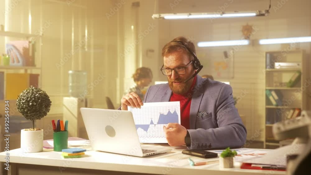 A middle aged man communicates via video conference using a laptop and a headset. A businessman in a blue jacket and a red turtleneck sits at a table in the office and demonstrates graphs and charts. A middle aged man communicates via video conference using a laptop and a headset. A businessman in a blue jacket and a red turtleneck sits at a table in the office and demonstrates graphs and charts.