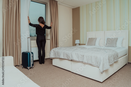 Young woman traveler opening the curtains and looking at the view from the window of a hotel room while on winter vacation, Travel lifestyle concept