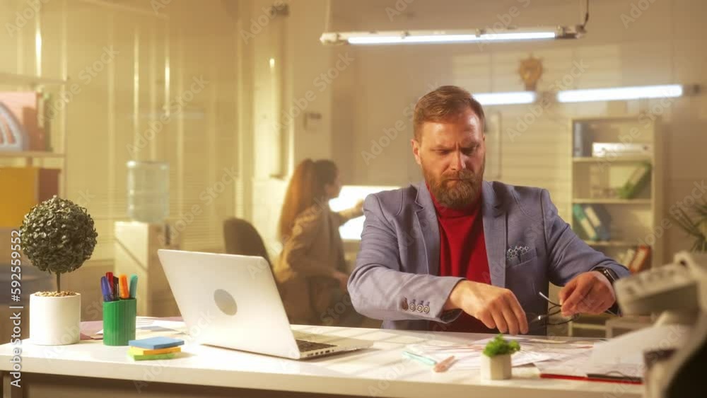 A middle aged man is typing on a laptop keyboard, suffering from vision problems and wearing glasses. A businessman with a beard in a blue jacket and a red turtleneck works in the office at the table. A middle aged man is typing on a laptop keyboard, suffering from vision problems and wearing glasses. A businessman with a beard in a blue jacket and a red turtleneck works in the office at the table.