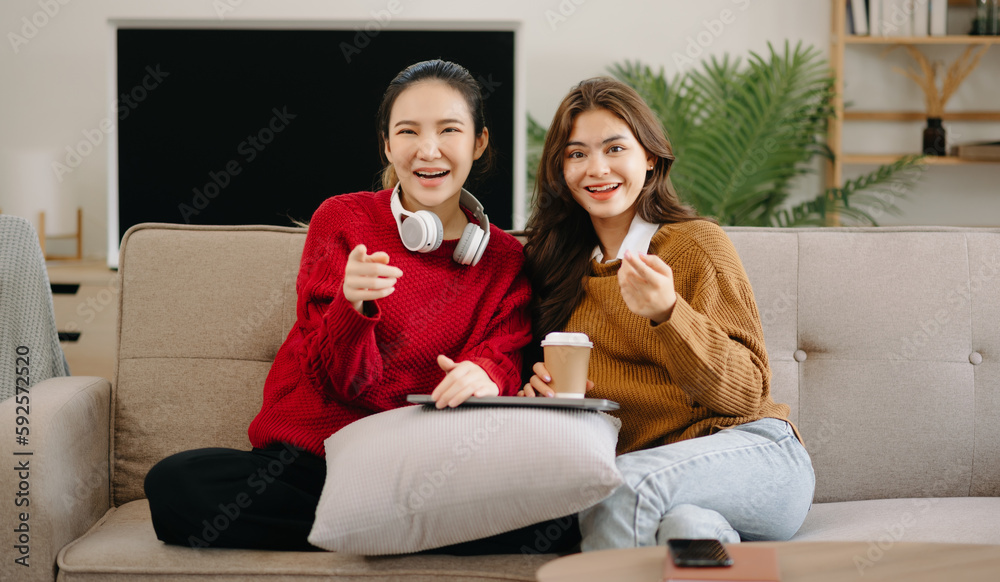 Obraz premium Two asian beauty smiling young women sitting on sofa Attractive casual girl feel happy and relax,having fun watch comedy video on TV in house. Activity lifestyles concept.