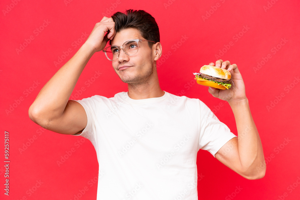 Young Caucasian man holding a burger isolated on red background having doubts and with confuse face expression Young Caucasian man holding a burger isolated on red background having doubts and with confuse face expression