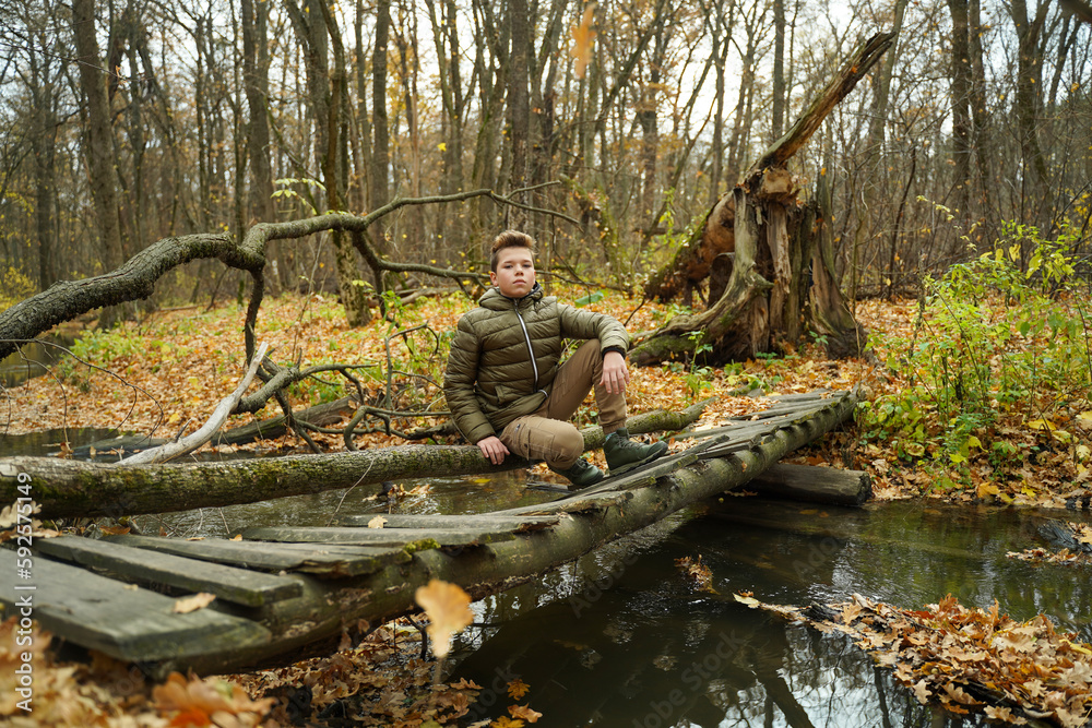 A young guy with a stylish hairstyle and fashionable clothes sits in the woods on a bridge. Autumn men's fashion
