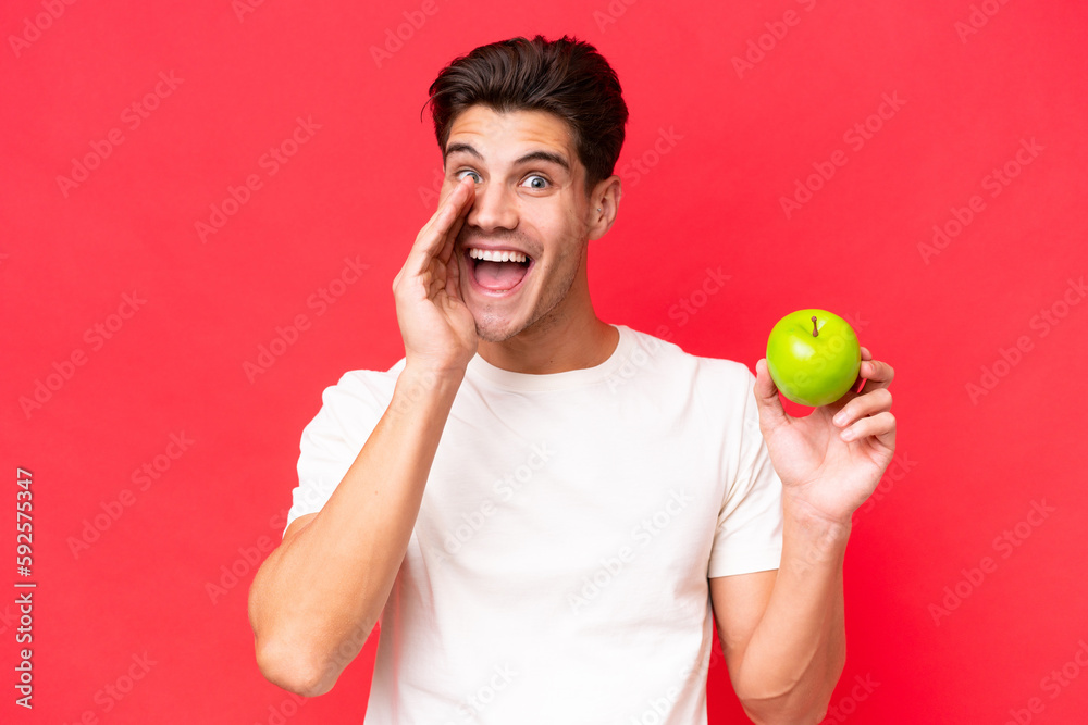 Young caucasian man with an apple isolated on red background shouting with mouth wide open