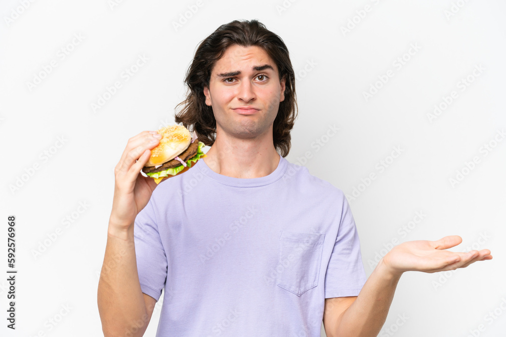 Young handsome man holding a burger isolated on white background making doubts gesture while lifting the shoulders Young handsome man holding a burger isolated on white background making doubts gesture while lifting the shoulders