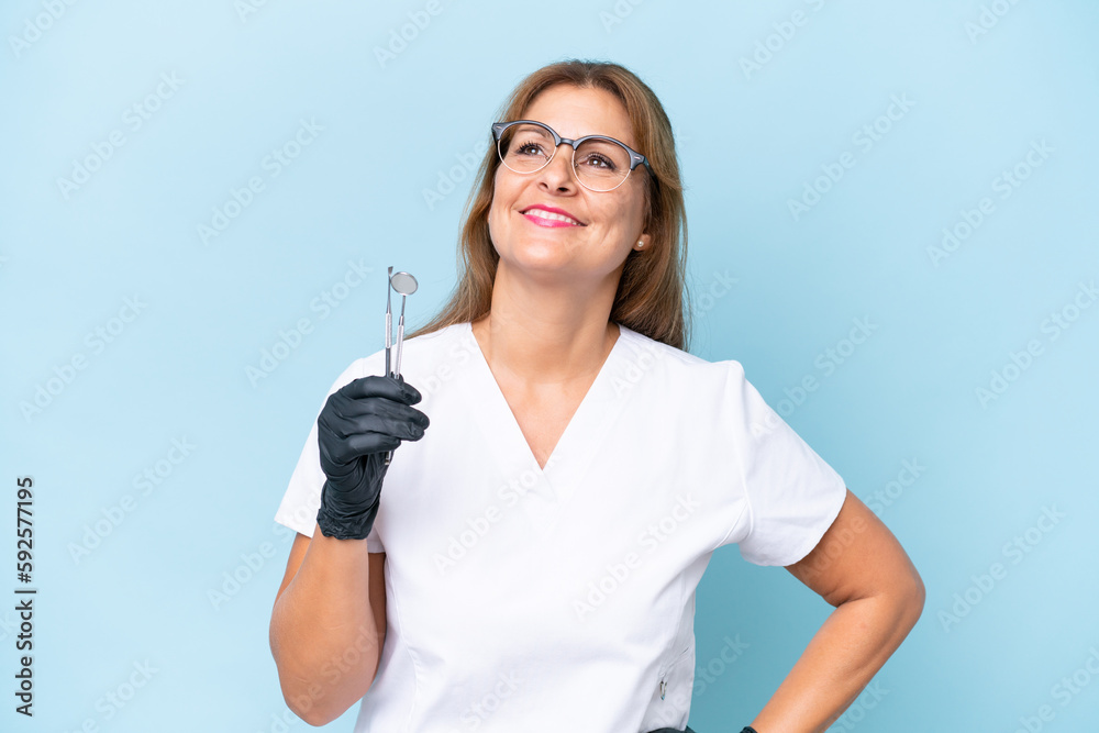 Middle-aged Dentist holding tools over isolated blue background posing with arms at hip and smiling Middle-aged Dentist holding tools over isolated blue background posing with arms at hip and smiling