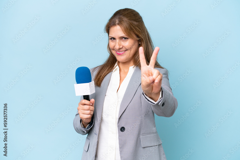 Middle-aged TV presenter woman over isolated blue background smiling and showing victory sign Middle-aged TV presenter woman over isolated blue background smiling and showing victory sign