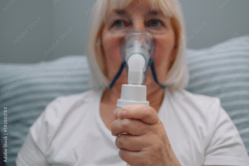 Sick upset caucasian senior woman with an inhaler. Unhealthy female doing inhalation at home, she use nebulizer and inhaler for the treatment, sitting on the couch at home, need treatment. Sick upset caucasian senior woman with an inhaler. Unhealthy female doing inhalation at home, she use nebulizer and inhaler for the treatment, sitting on the couch at home, need treatment.