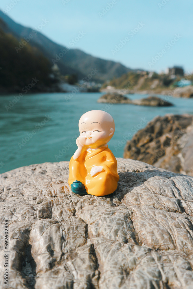 A small buddha on the rocks near the river against the backdrop of the mountains. Figurine of a monk in orange robes on the banks of the Ganges. A small buddha on the rocks near the river against the backdrop of the mountains. Figurine of a monk in orange robes on the banks of the Ganges.