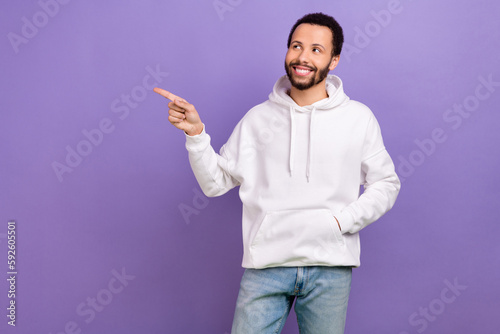Portrait of cheerful guy with brunet beard wear white pullover directing look at discount empty space isolated on violet color background