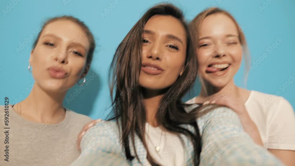 Three young smiling hipster women in summer clothes. Girls taking selfie self portrait photos or video on smartphone. Models posing in the studio near blue wall Three young smiling hipster women in summer clothes. Girls taking selfie self portrait photos or video on smartphone. Models posing in the studio near blue wall