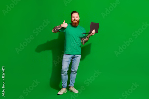 Full length photo of cool confident man wear t-shirt showing thumb up reading modern gadget isolated green color background © deagreez