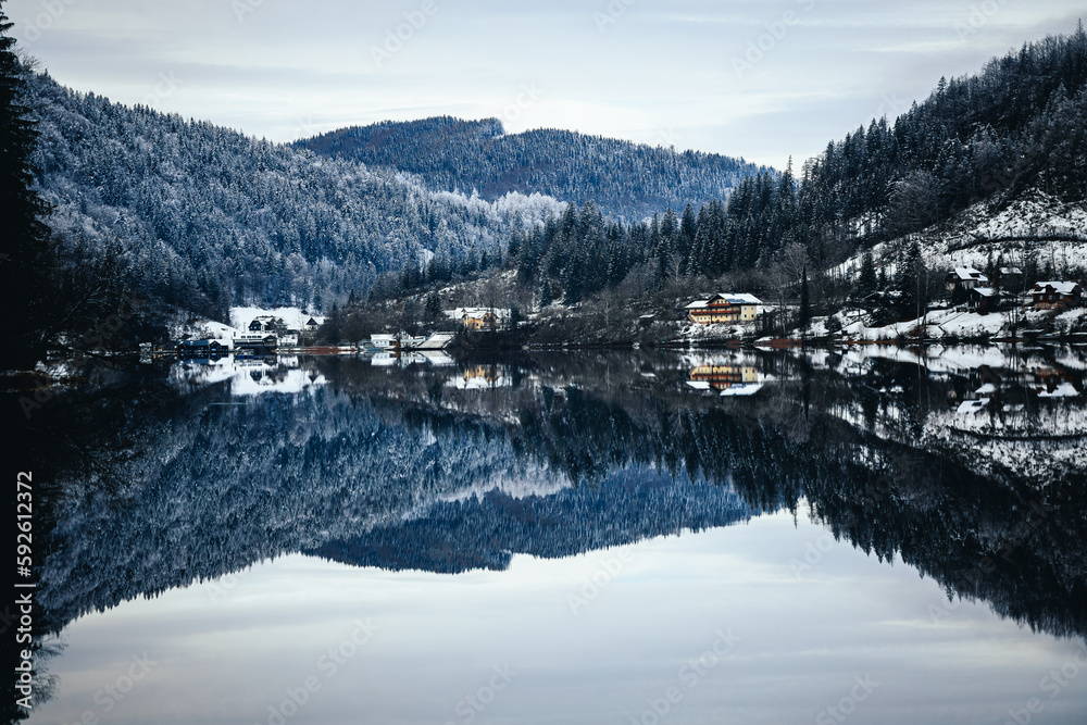 Fototapeta premium Houses on the lakeside against a mountain covered with trees in winter with reflection in water
