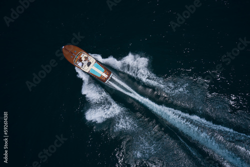 Luxurious wooden boat with people moving fast on dark water top view. Classic Italian wooden boat fast moving aerial view. Top view of a wooden powerful motor boat.