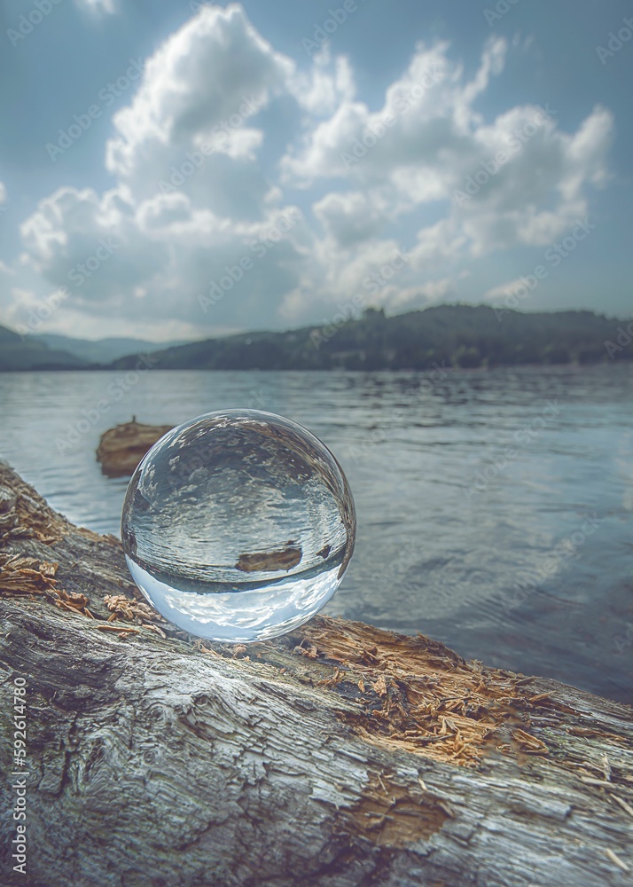 Vertical shot of a crystal ball on a fallen tree log, reflecting a tranquil lake on a cloudy day Vertical shot of a crystal ball on a fallen tree log, reflecting a tranquil lake on a cloudy day