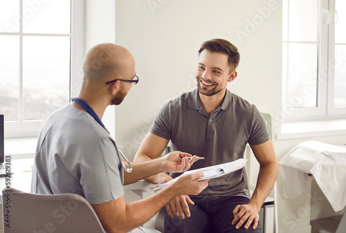 Portrait of a happy smiling young man patient in medical office listening to doctor holding report file with appointment and giving consultation during medical examination in clinic. photo