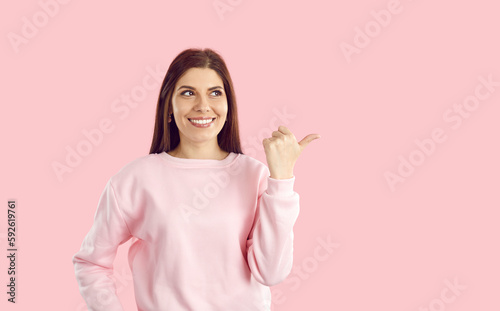 Cheerful pretty young woman smiling and pointing finger at copy space for your great offer or promotional banner with useful information dressed in casual style, stands on pastel pink background