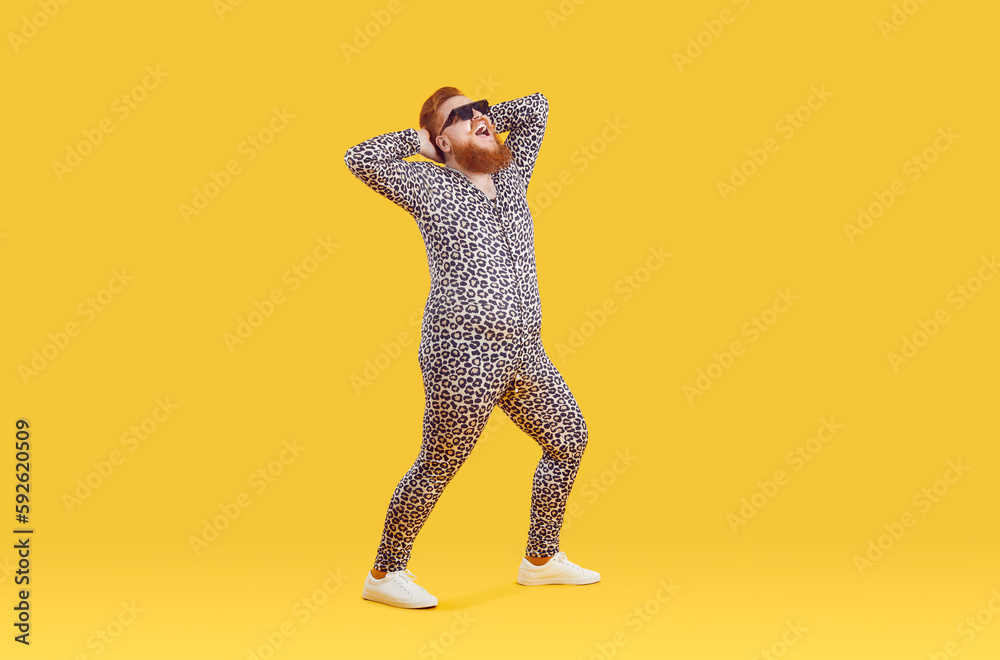 Fototapeta premium Funny cheerful fat guy in crazy animal print PJs having fun in modern fashion studio. Happy excited fat man wearing comfortable leopard pajamas and sunglasses posing isolated on yellow background