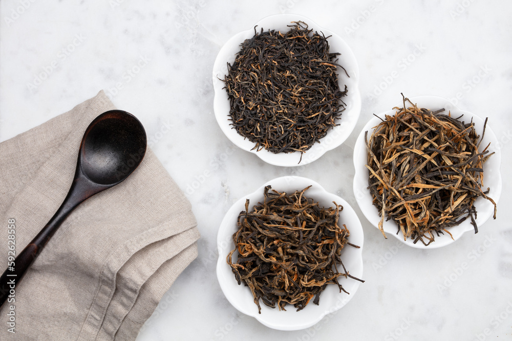 Fototapeta premium Dry tea in a white bowls and empty wooden spoon on a white marble background. Dried tea leaves. Tree white ceramic bowls with dried tea leaves, close up. Top view.