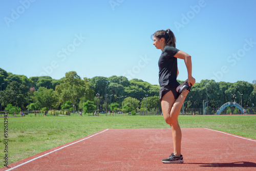 side view of young latin woman standing on running track stretching femoral muscle leg.
