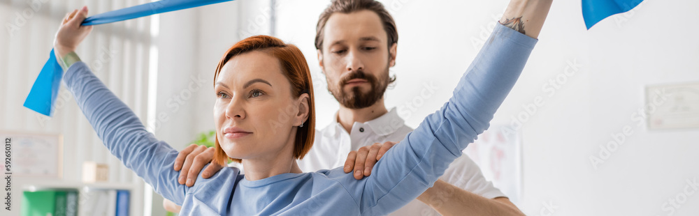 physiotherapist assisting redhead woman working out with resistance band in rehabilitation center, banner. physiotherapist assisting redhead woman working out with resistance band in rehabilitation center, banner.