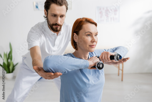 rehabilitologist assisting redhead woman working out with dumbbells in recovery center. photo