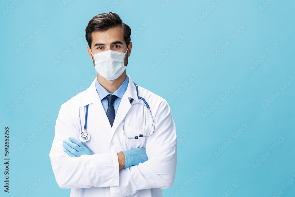 A male doctor in a white coat and a medical mask stands with his hands and looks at the camera on a blue isolated background, copy space, space for text A male doctor in a white coat and a medical mask stands with his hands and looks at the camera on a blue isolated background, copy space, space for text