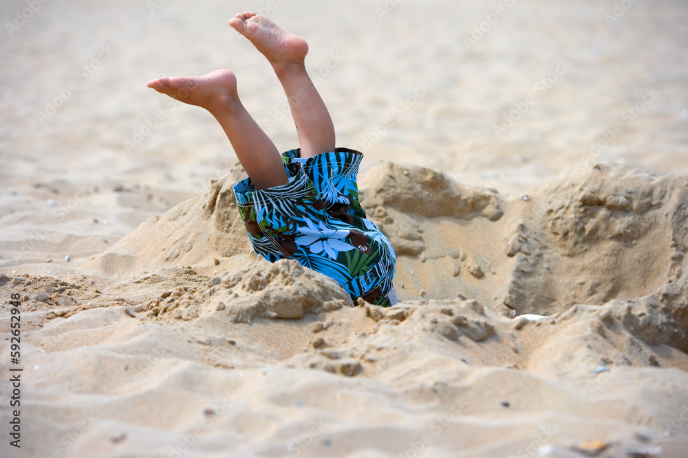 Child has fallen in a pit on the beach, only his legs are seen  - obrazy, fototapety, plakaty 