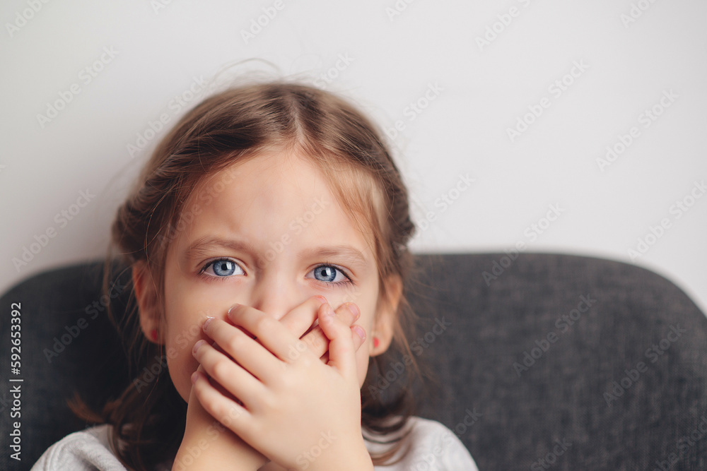 Little girl covers mouth with hand. Sad offended child girl sitting at sofa at home. Child abuse. Little girl covers mouth with hand. Sad offended child girl sitting at sofa at home. Child abuse.