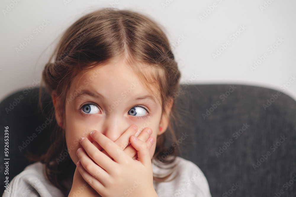Little girl covers mouth with hand. Sad offended child girl sitting at sofa at home. Child abuse. Little girl covers mouth with hand. Sad offended child girl sitting at sofa at home. Child abuse.