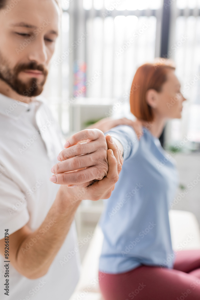bearded physiotherapist stretching painful arm of blurred woman during diagnostics in consulting room.