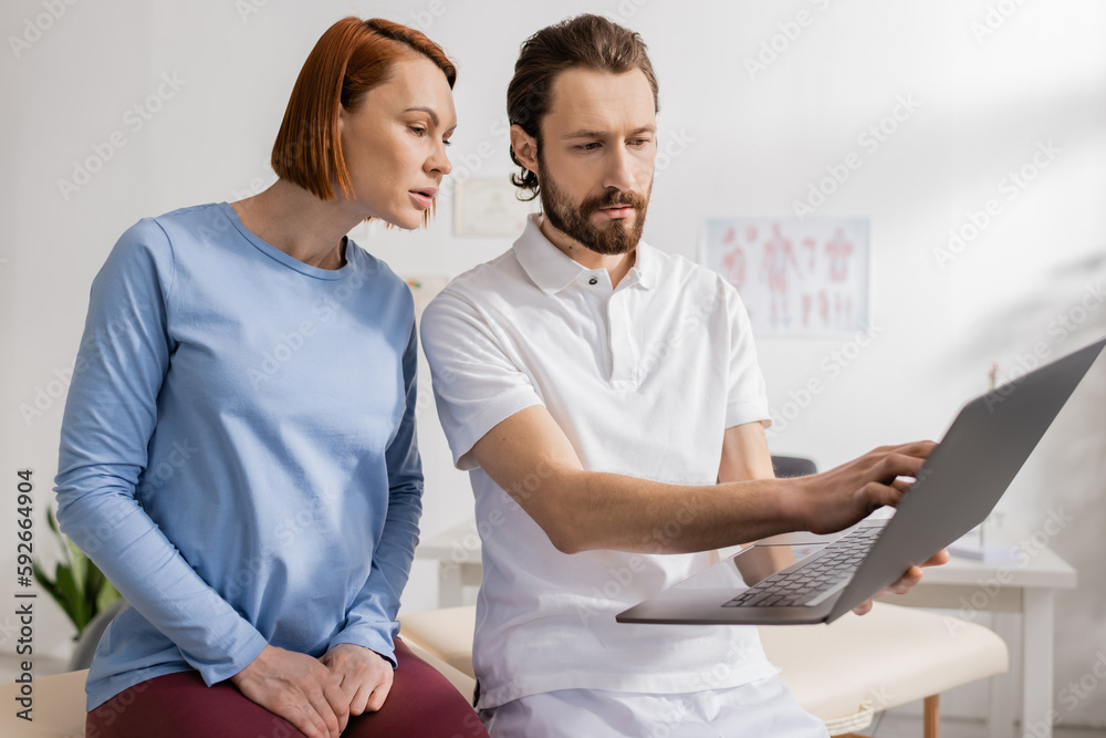 bearded manual therapist showing laptop to redhead woman during consultation in rehabilitation center. bearded manual therapist showing laptop to redhead woman during consultation in rehabilitation center.