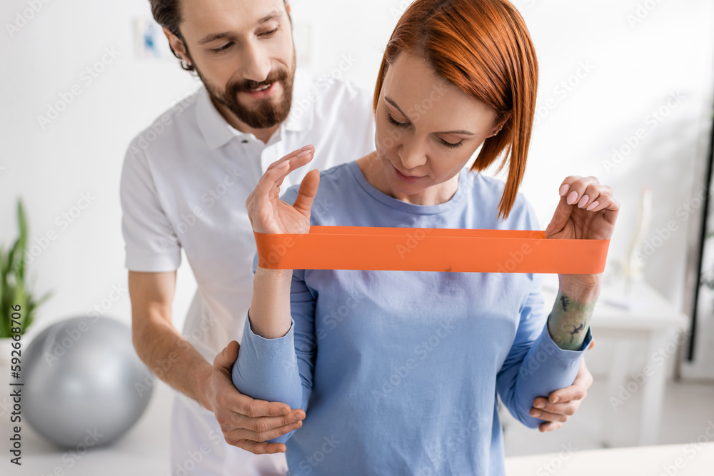Fototapeta premium redhead woman training arms with resistance band near bearded physiotherapist in rehab center.