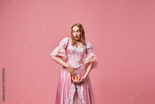 Portrait of young beautiful woman  queen wearing dress and holding grapefruit down with surprised face over pink studio background. Femininity
