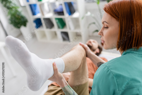 redhead physiotherapist flexing leg of man during recovery therapy in consulting room.