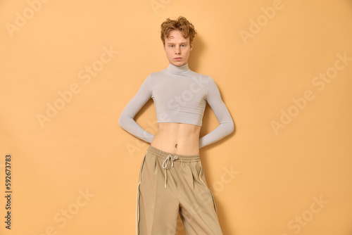 Cocky caucasian gay man with short curly red hair wearing trendy beige crop top and pants standing with bare belly near yellow wall with hands behind his back and looking at camera with confidence.