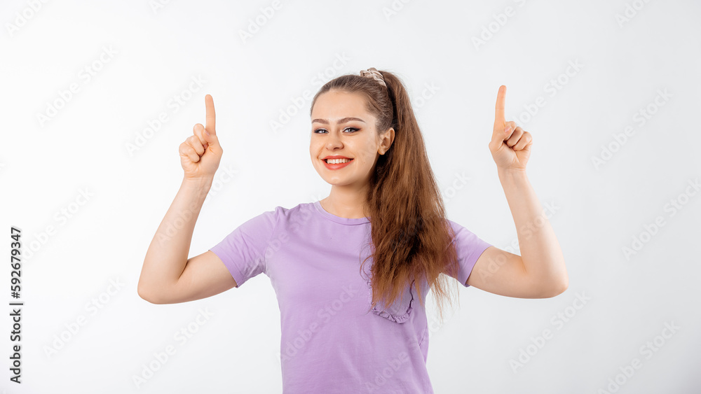 Happy young woman with long hair pointing fingers up and smiling, showing advertisement on top, product promotion, promo offer, standing in casual t shirt over white background Happy young woman with long hair pointing fingers up and smiling, showing advertisement on top, product promotion, promo offer, standing in casual t shirt over white background