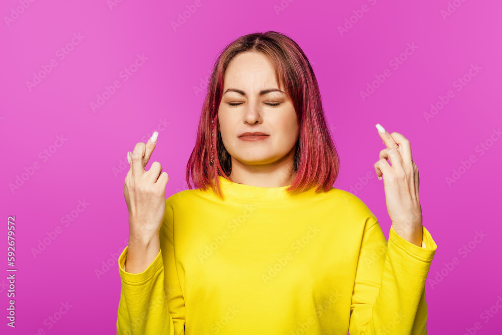 Hopeful young woman with pink hair close eyes and holds fingers crossed, while making wish, standing in yellow t-shirt on pink background Hopeful young woman with pink hair close eyes and holds fingers crossed, while making wish, standing in yellow t-shirt on pink background