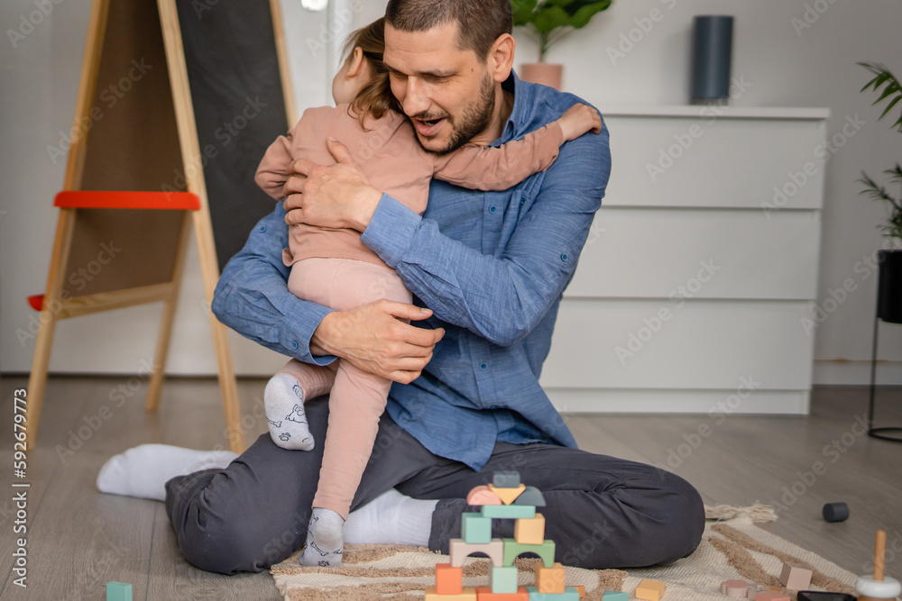 Fototapeta premium One toddler girl caucasian hugging her father while they playing at home on the floor with wooden toys, modern parenting lifestyle love emotion happy family concept 