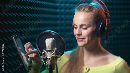 Girl in recording studio with mic scrolling digital tablet for text or notes, over acoustic absorber panel background