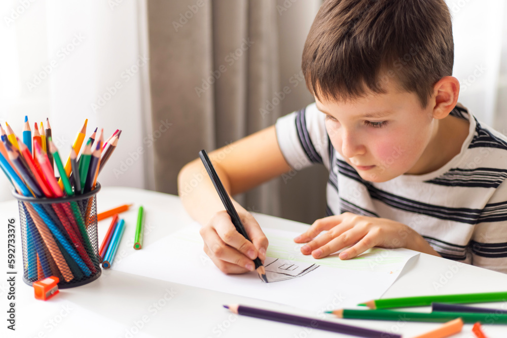 A child boy draws on white paper with colored pencils while sitting at a table A child boy draws on white paper with colored pencils while sitting at a table