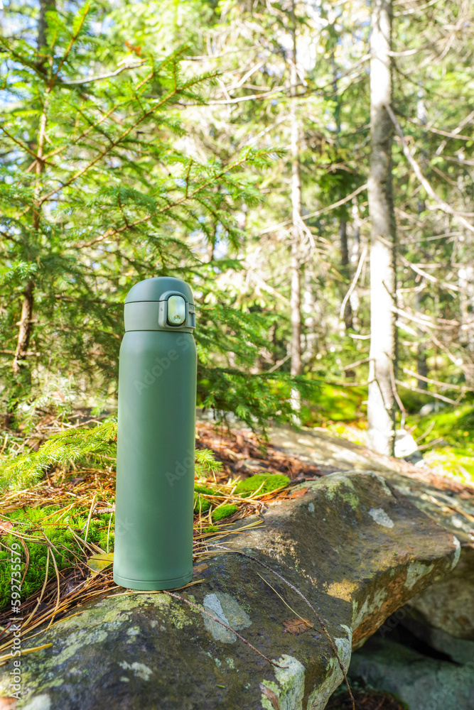 Green thermos on the stone. Thermal bottle on the background of the forest. Sunny day in the forest. Green thermos on the stone. Thermal bottle on the background of the forest. Sunny day in the forest.