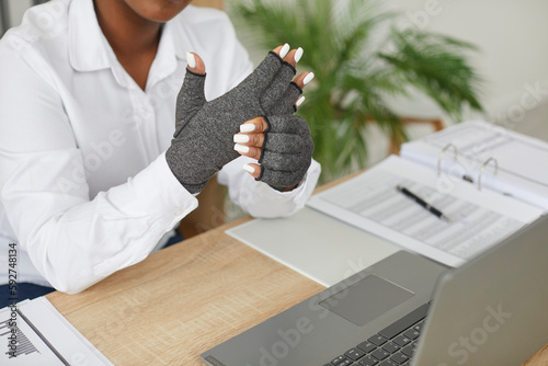 Foto Young African American business woman with rheumatoid arthritis wears compression gloves to help relieve hand pain while typing on a laptop computer keyboard at work in the office