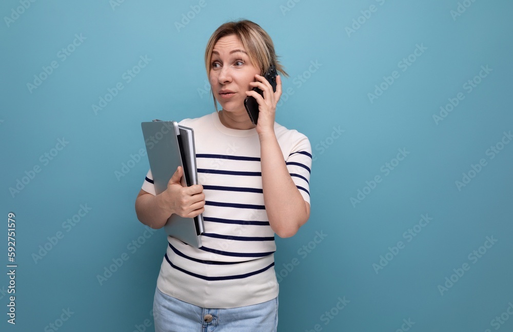 confident smiling blond girl talking on a smartphone holding a laptop in her hands on a blue background with copy space confident smiling blond girl talking on a smartphone holding a laptop in her hands on a blue background with copy space
