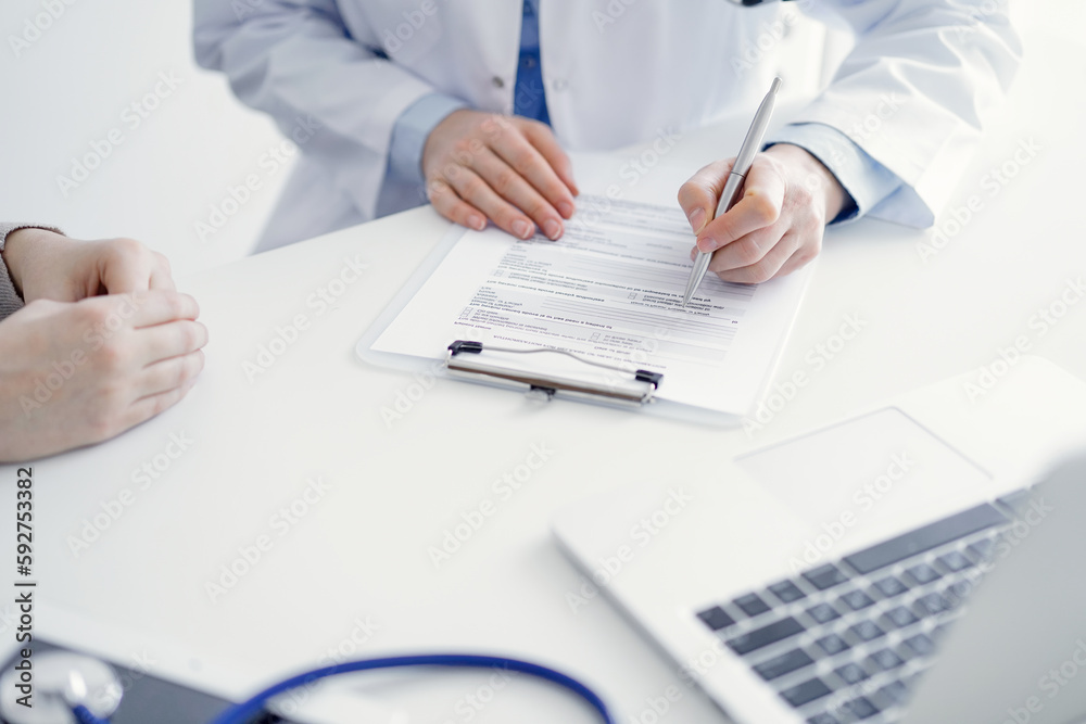 Doctor and patient sitting at the table in clinic. The focus is on female physician's hands filling up the medication history record form or checklist, close up. Medicine concept Doctor and patient sitting at the table in clinic. The focus is on female physician's hands filling up the medication history record form or checklist, close up. Medicine concept