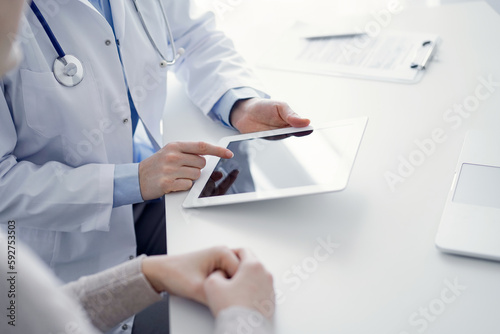 Doctor and patient sitting at the table in clinic. The focus is on female physician's hands using tablet computer, close up. Medicine and healthcare concept