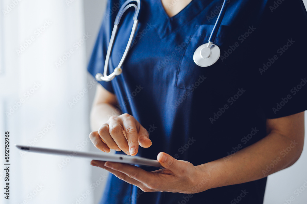 Doctor woman using tablet computer while standing near panorama window in clinic, close up. Physician or surgeon at work. Medicine concept Doctor woman using tablet computer while standing near panorama window in clinic, close up. Physician or surgeon at work. Medicine concept